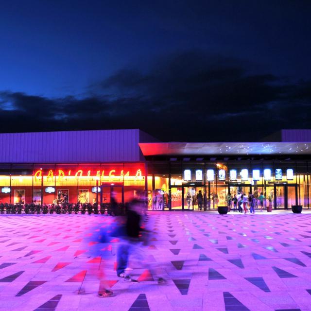 Personne marchant sur l'esplanade des ruthènes, avec un éclairage violet, devant la vitrine du Cinéma de Rodez