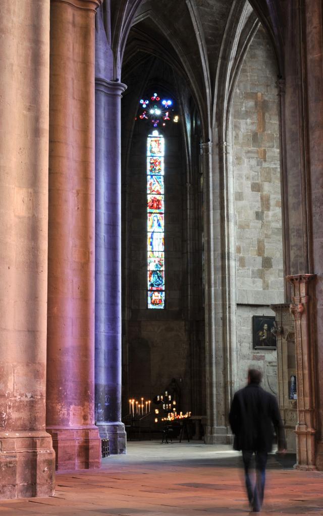 Personne qui déambule aux pieds des piliers à l'intérieur de la cathédrale de Rodez illuminée par les vitraux et les cierges allumés