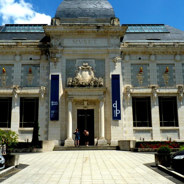 Façade principale du musée des Beaux-Arts Denys-Puech à Rodez, avec ses chiens jaunes sculptés et médaillons, devant un parking