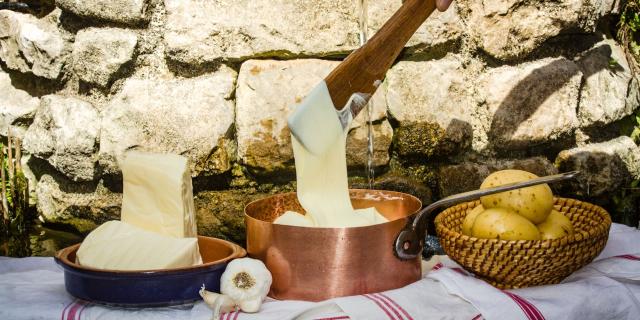 Devant un mur de pierres, sur des torchons, un plat en terre cuite avec deux morceaux de tomme, une casserole avec de l'aligot filant depuis une cuilllère en bois, et un plat en osier avec des pommes de terre, de l'ail