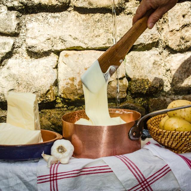 Devant un mur de pierres, sur des torchons, un plat en terre cuite avec deux morceaux de tomme, une casserole avec de l'aligot filant depuis une cuilllère en bois, et un plat en osier avec des pommes de terre, de l'ail