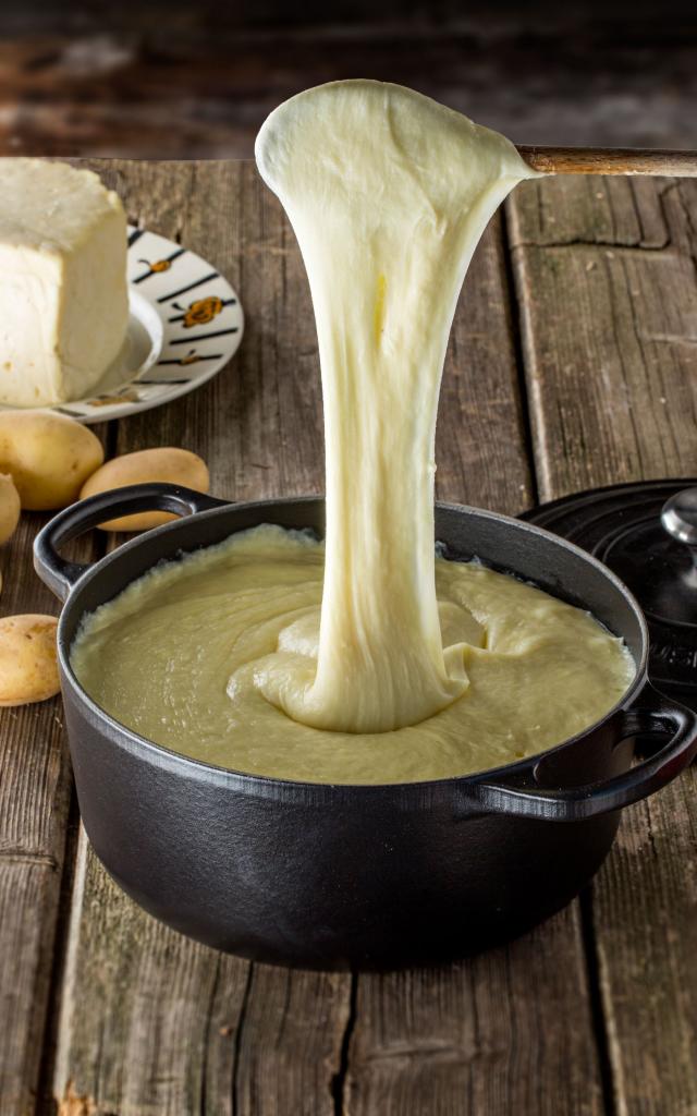 Sur une table en bois, plat d'aligot dans une marmite filant depuis une cuillère en bois. Pommes de terre et tomme en arrière plan