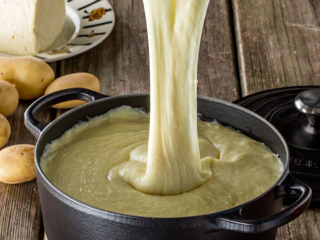 sur une table en bois, plat d'aligot dans une marmite filant depuis une cuillère en bois. Pommes de terre et tomme en arrière plan