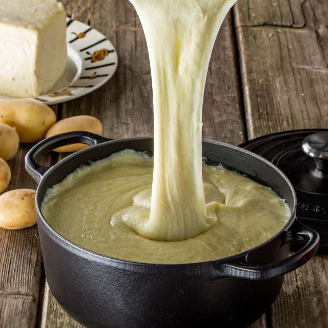 Sur une table en bois, plat d'aligot dans une marmite filant depuis une cuillère en bois. Pommes de terre et tomme en arrière plan