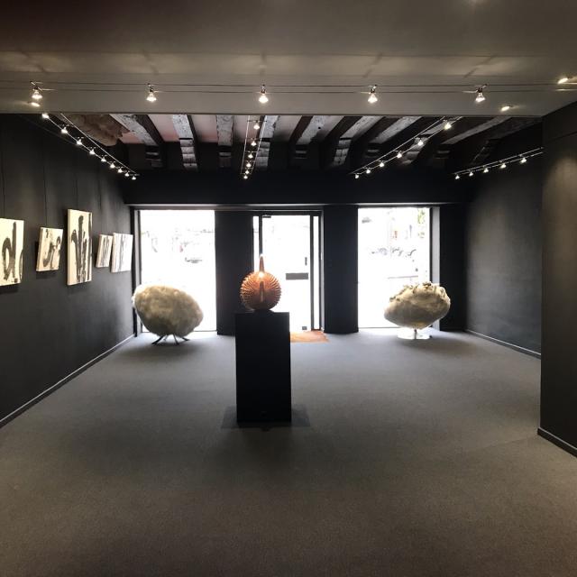 Salle d'exposition de la Galerie Annotiau aux murs noirs avec des cadres noir et blanc accrochés, ainsi que deux fauteuils face à la baie vitrée et une sculpture au centre