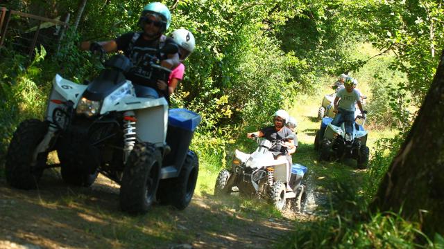 Quatre quad avec une ou deux personnes, casquées, se suivent à la file dans un chemin entre les arbres