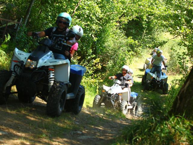 Quatre quad avec une ou deux personnes, casquées, se suivent à la file dans un chemin entre les arbres