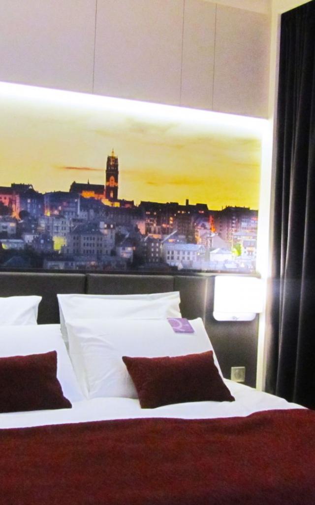 Lit double de l'Hôtel Mercure avec coussins blancs et par-dessus bordeaux avec une tête de lit en cuir noir, au-dessous un tableau panoramique de la ville de Rodez et sa cathédrale au coucher du soleil