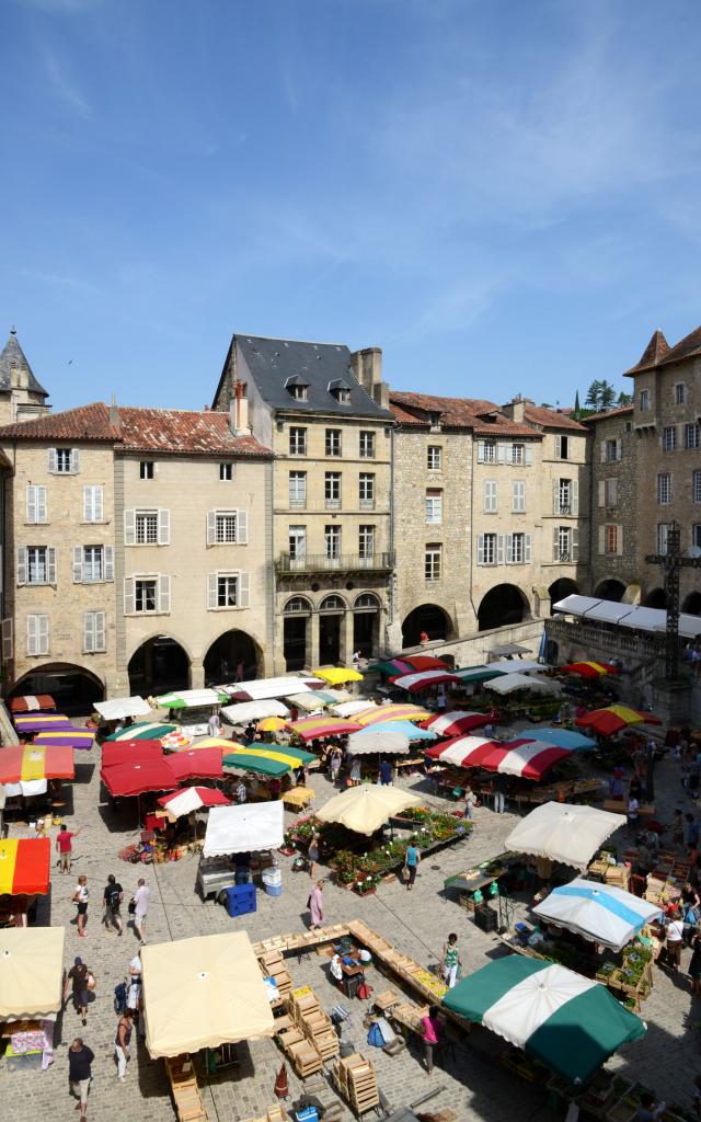 Personnes au marché de Villefranche de Rouergue avec des marchands ambulants sous leurs parasols, au cœur d'une place pavée avec des arcades