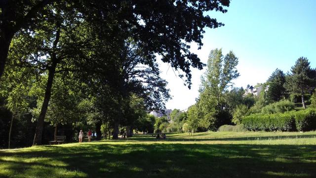 Personne qui se baladent sur le chemin piétonnier au bord de l'espace vert arboré à Layoule où des gens sont assis dans l'herbe