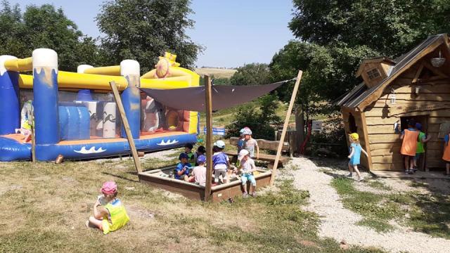 Groupe d'enfants avec casquettes jouant dans un bac à sable carré en bois, à côté d'une cabane en bois et de jeux gonflables, dans un espace vert