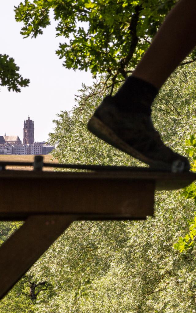 Paire de jambes en équilibre sur une corde dans les arbres lors d'un accrobranche, descendant d'un palier fixe en bois, avec une vue lointaine sur la cathédrale de Rodez