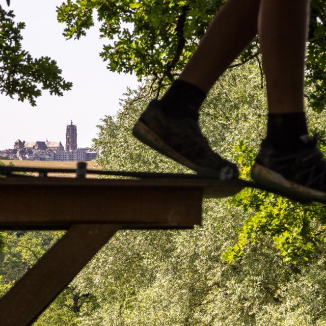 Paire de jambes en équilibre sur une corde dans les arbres lors d'un accrobranche, descendant d'un palier fixe en bois, avec une vue lointaine sur la cathédrale de Rodez