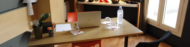 Bureau de l'hôtel Ibis avec table, chaises, ordinateur portable, bouteille d'eau et verres, cafetière et dosettes