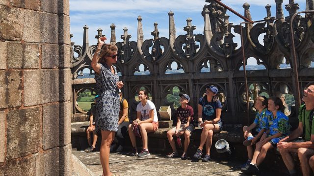 Groupe assis sur des rebords en pierre au sommet de la cathédrale, face à la guide-conférencière et dos aux détails sculptés du sommet