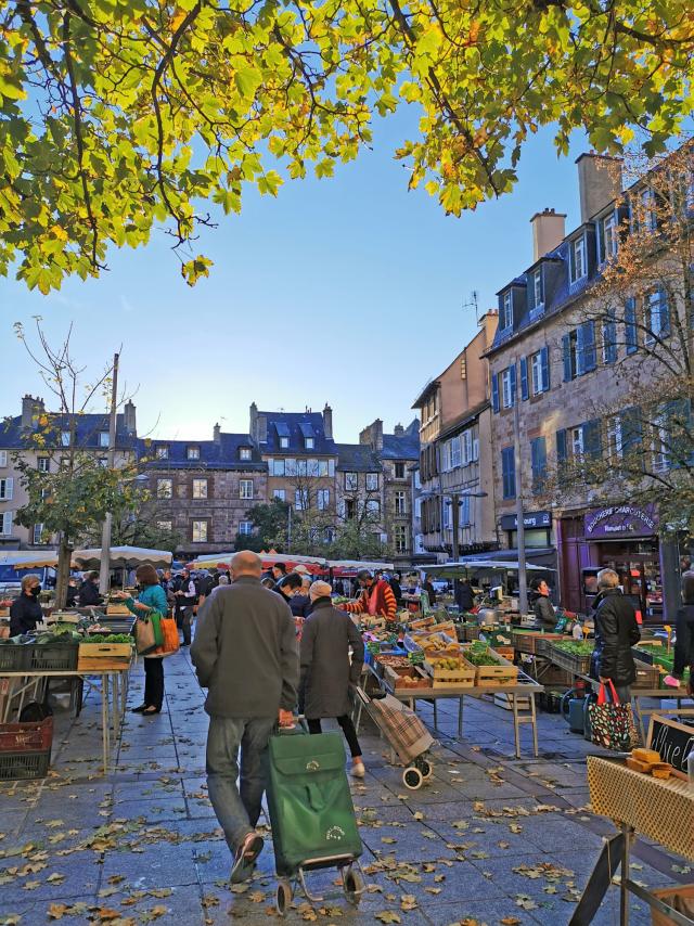 Personnes avec caddie marchant sur des feuilles mortes entre les étals de légumes, sur la place du marché devant les maisons typiques de Rodez