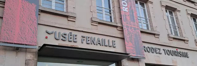 Devanture vitrée surmontée des enseignes Rodez Tourisme et musée Fenaille, agrémentées de bandeaux verticaux courant le long du bâtiment, avec le nom de l'enseigne et une photo associée, couleur rouge